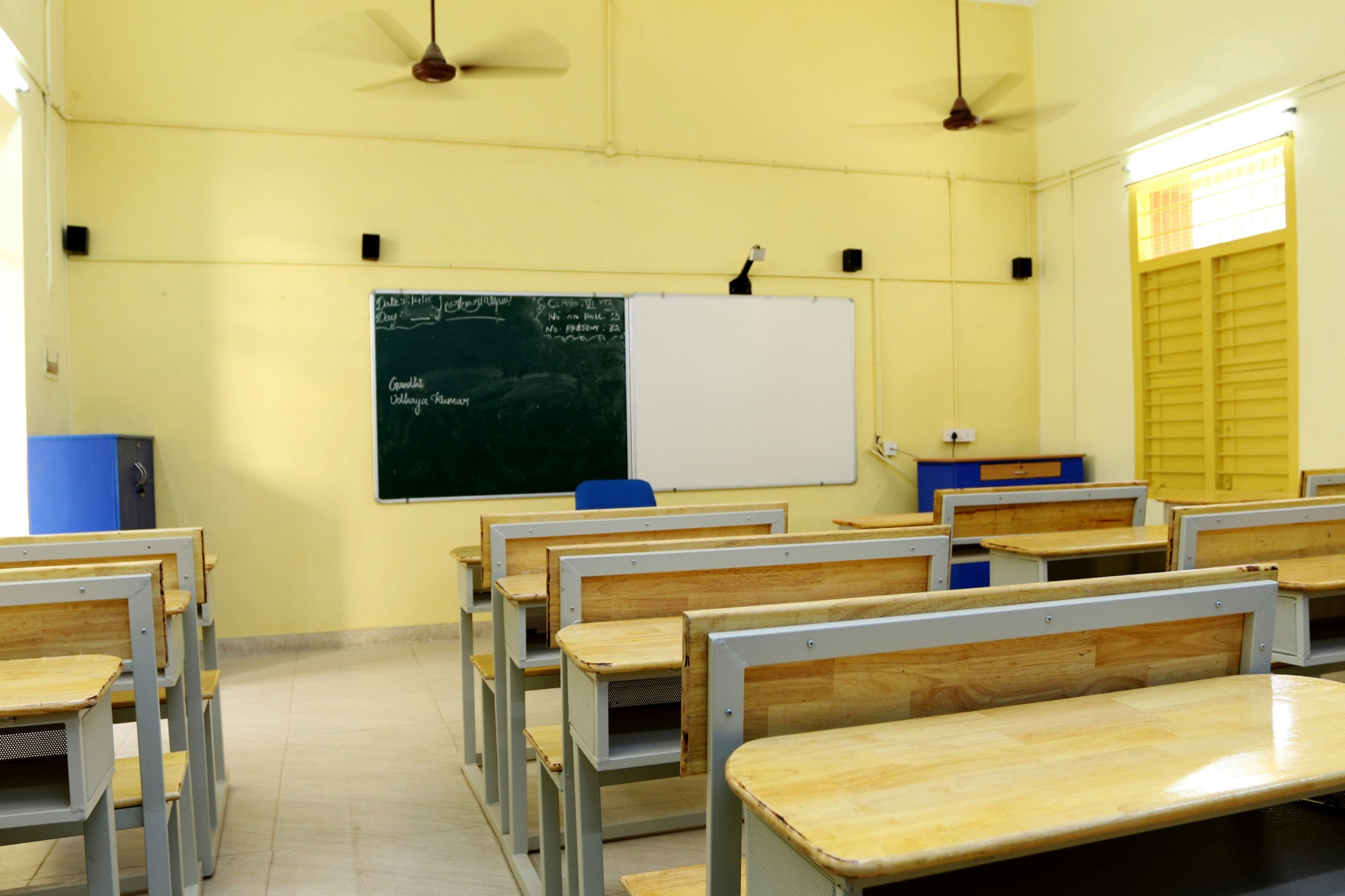 An empty classroom with pale yellow walls and rows of wooden bench desks, a green chalkboard beside a whiteboard at the front, ceiling fans overhead and a shuttered window to one side.