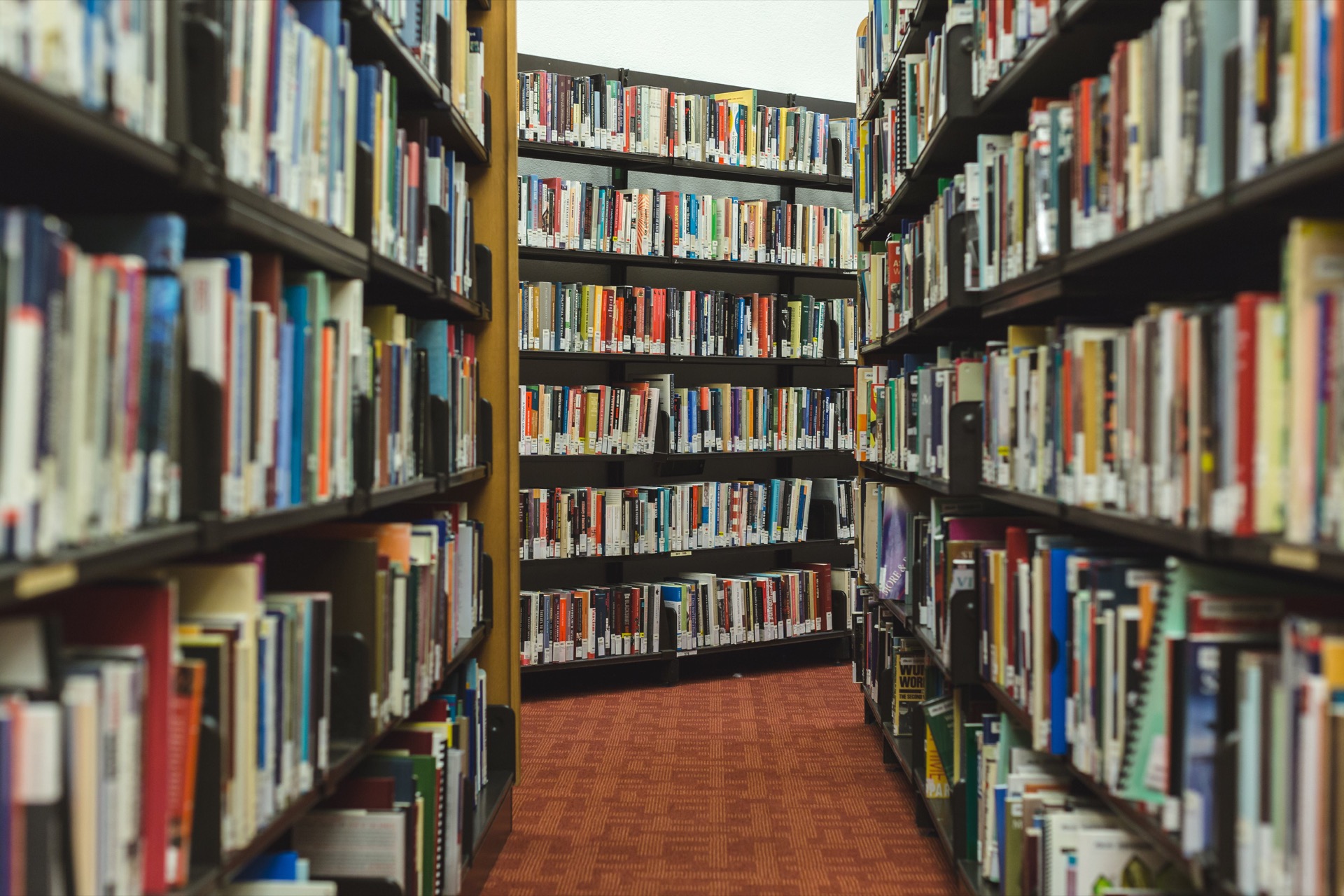 Looking down a narrow aisle between two tall library shelves packed tight with books, a red carpet running away to a further shelf at the end.