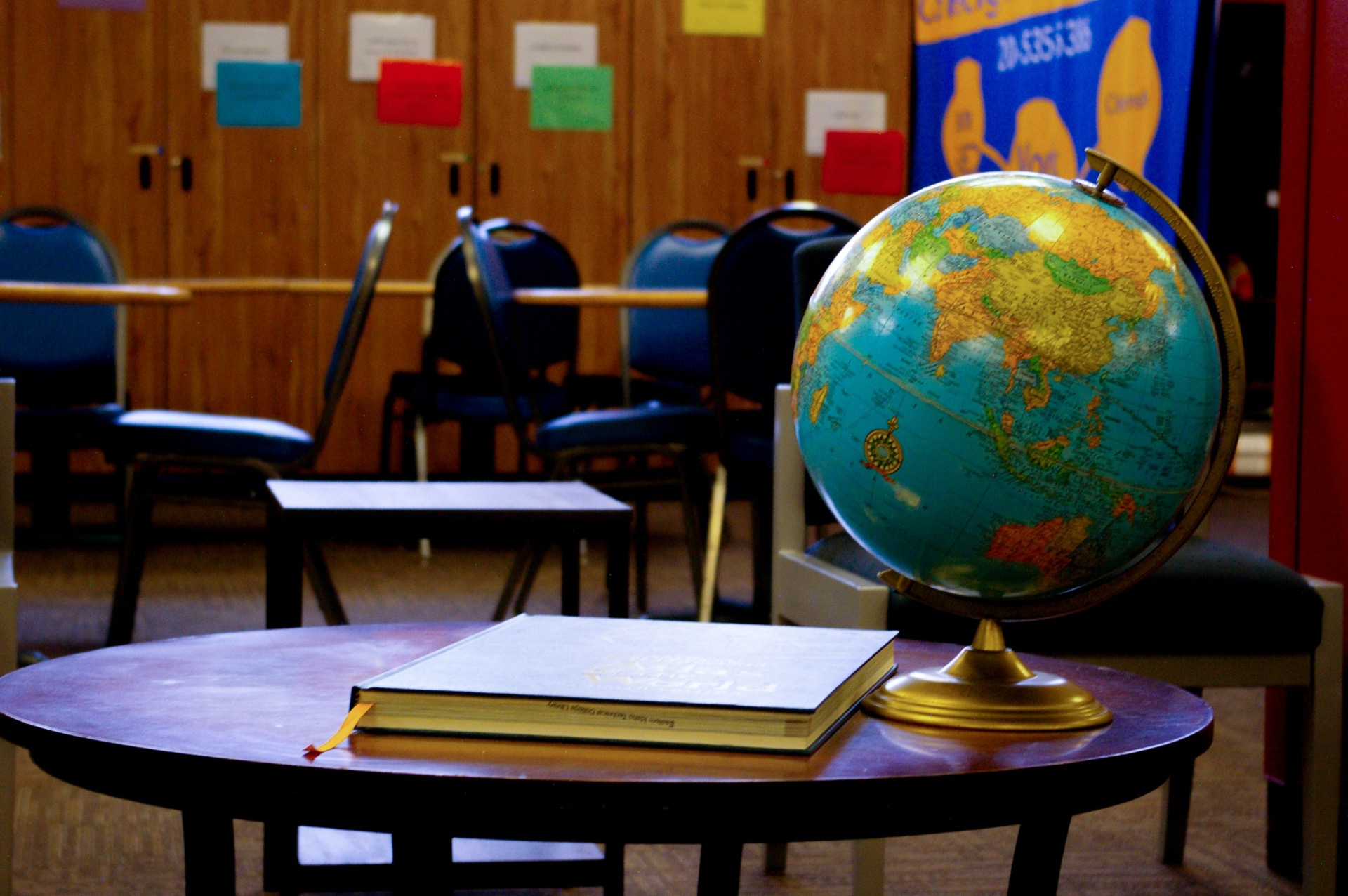 A world globe on a brass stand beside a large closed reference book on a round wooden table, with stacked chairs and a noticeboard of coloured cards behind.