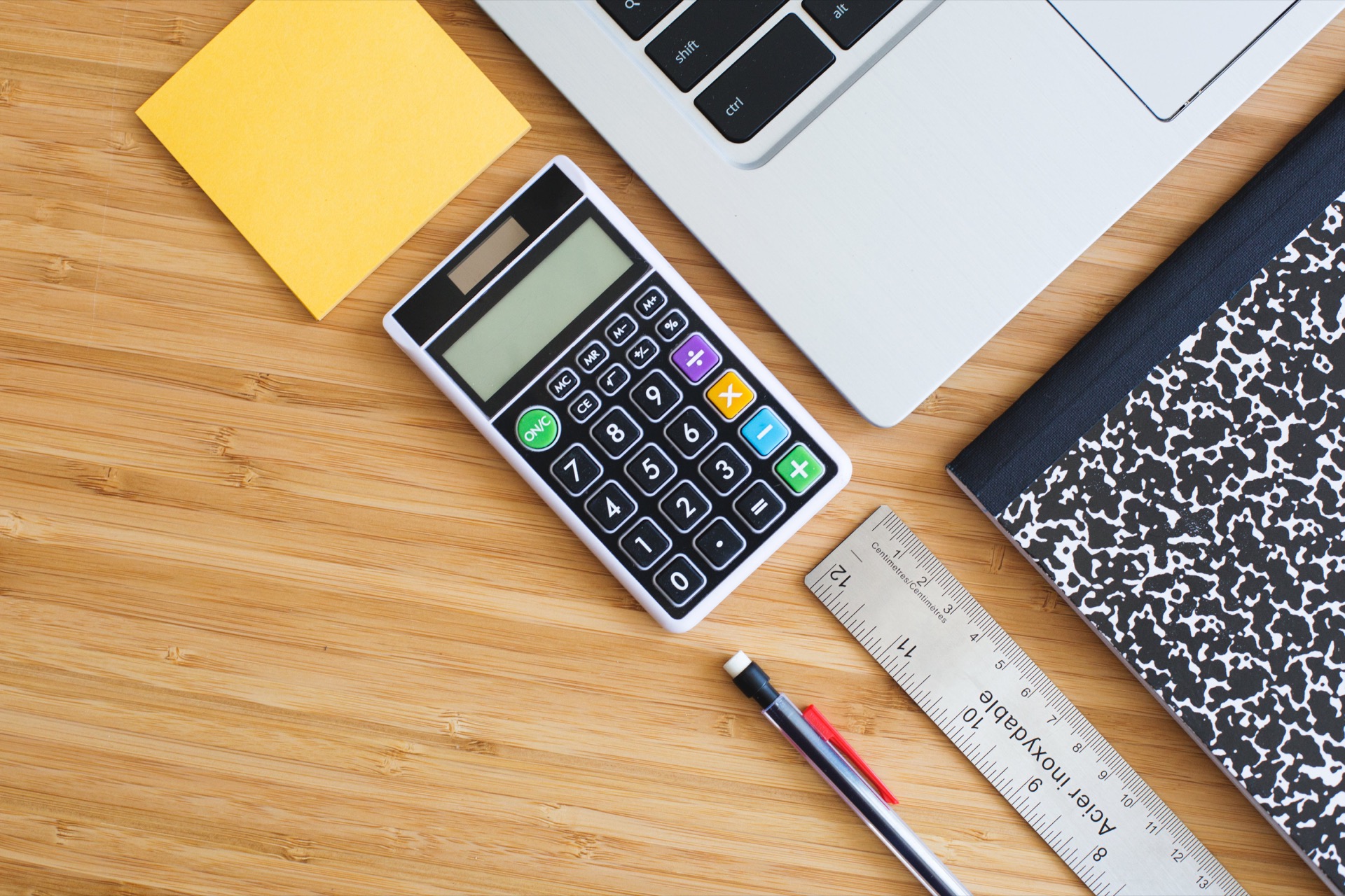 Overhead view of a desk: a pocket calculator, the corner of a laptop keyboard, a pad of yellow sticky notes, a steel ruler, a pen and a marbled composition notebook on pale bamboo.