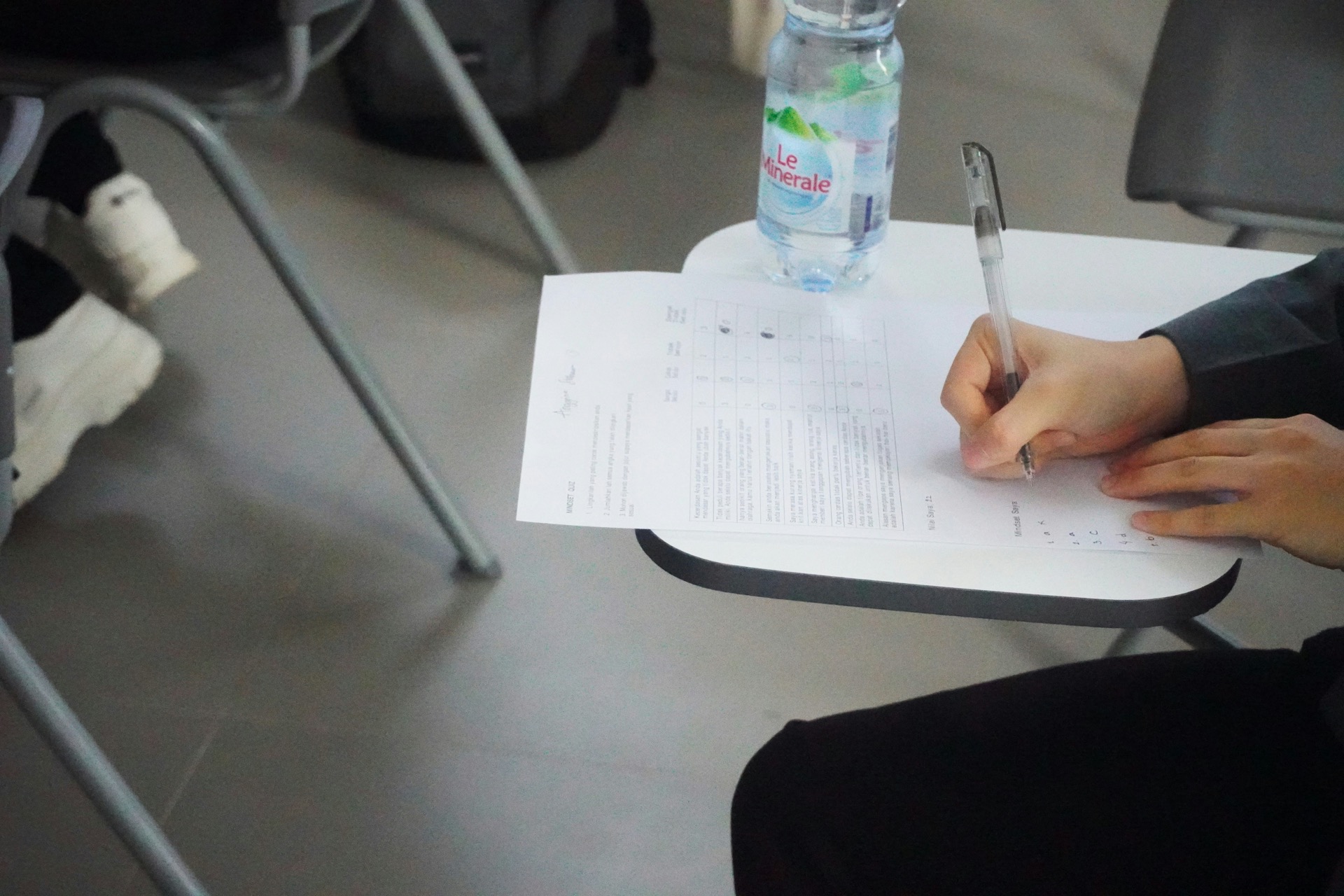 A student writes on a paper questionnaire balanced on the fold-out arm of a chair, a bottle of water on the surface beside them; the sheet shows a grid of response options down one side.