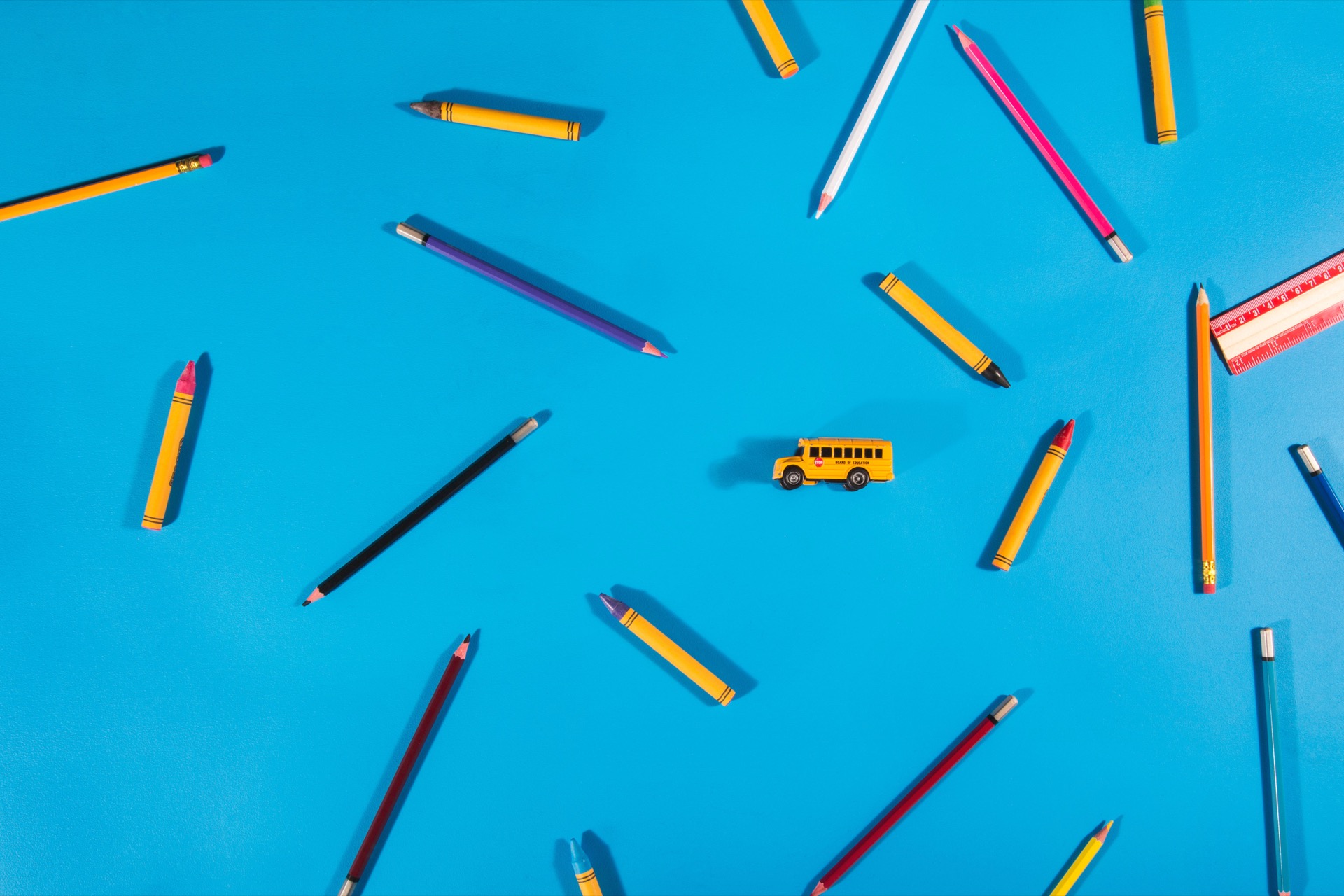 Overhead view on a bright blue background of crayons, coloured pencils and a ruler scattered outwards from a miniature yellow school bus at the centre.