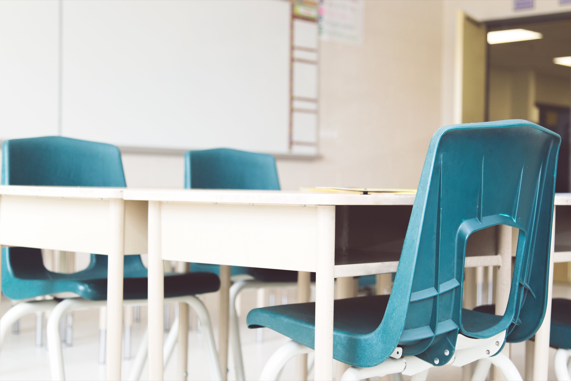 An empty classroom with teal moulded-plastic chairs pushed under cream desks, a blank whiteboard on the wall behind and a door standing open to the corridor.