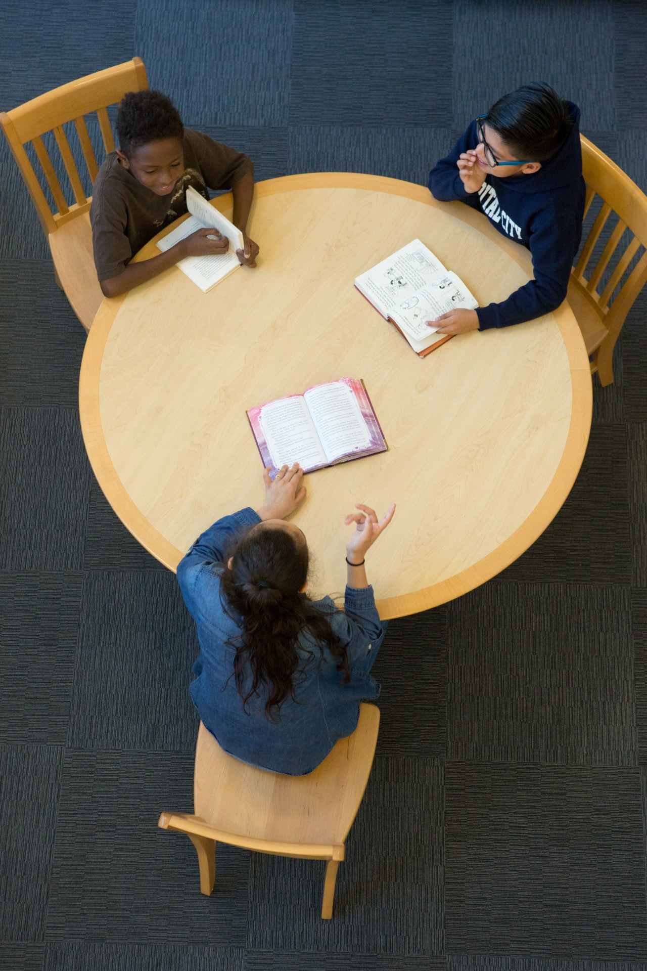 Seen from above, three children read at a round library table, two with books open in front of them and one holding a book up to read.