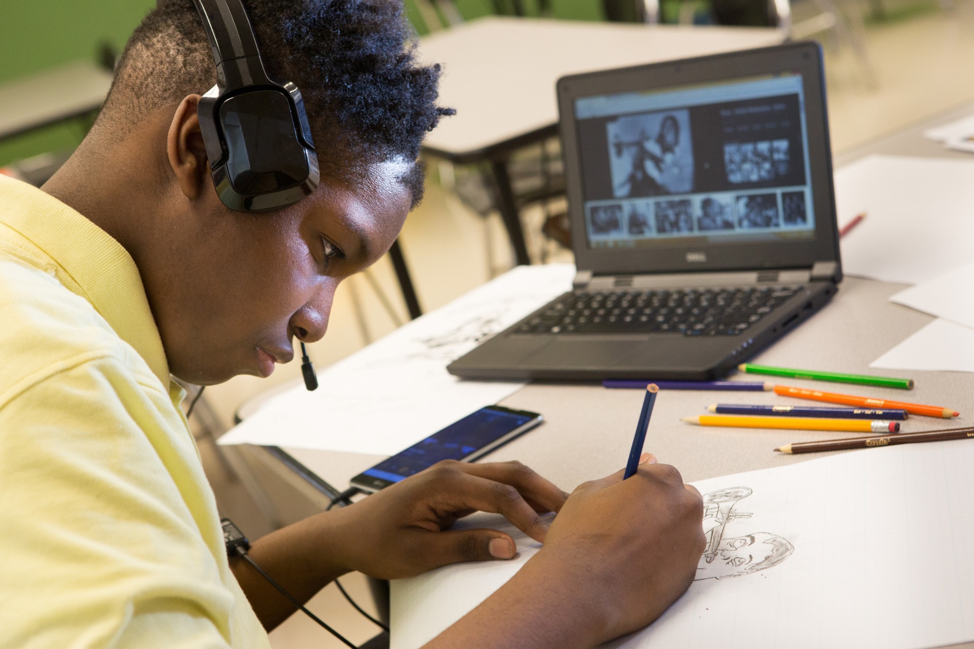 A student wearing over-ear headphones draws in pencil at a desk, working from a photograph displayed on the laptop screen beside them, with coloured pencils scattered across the table.