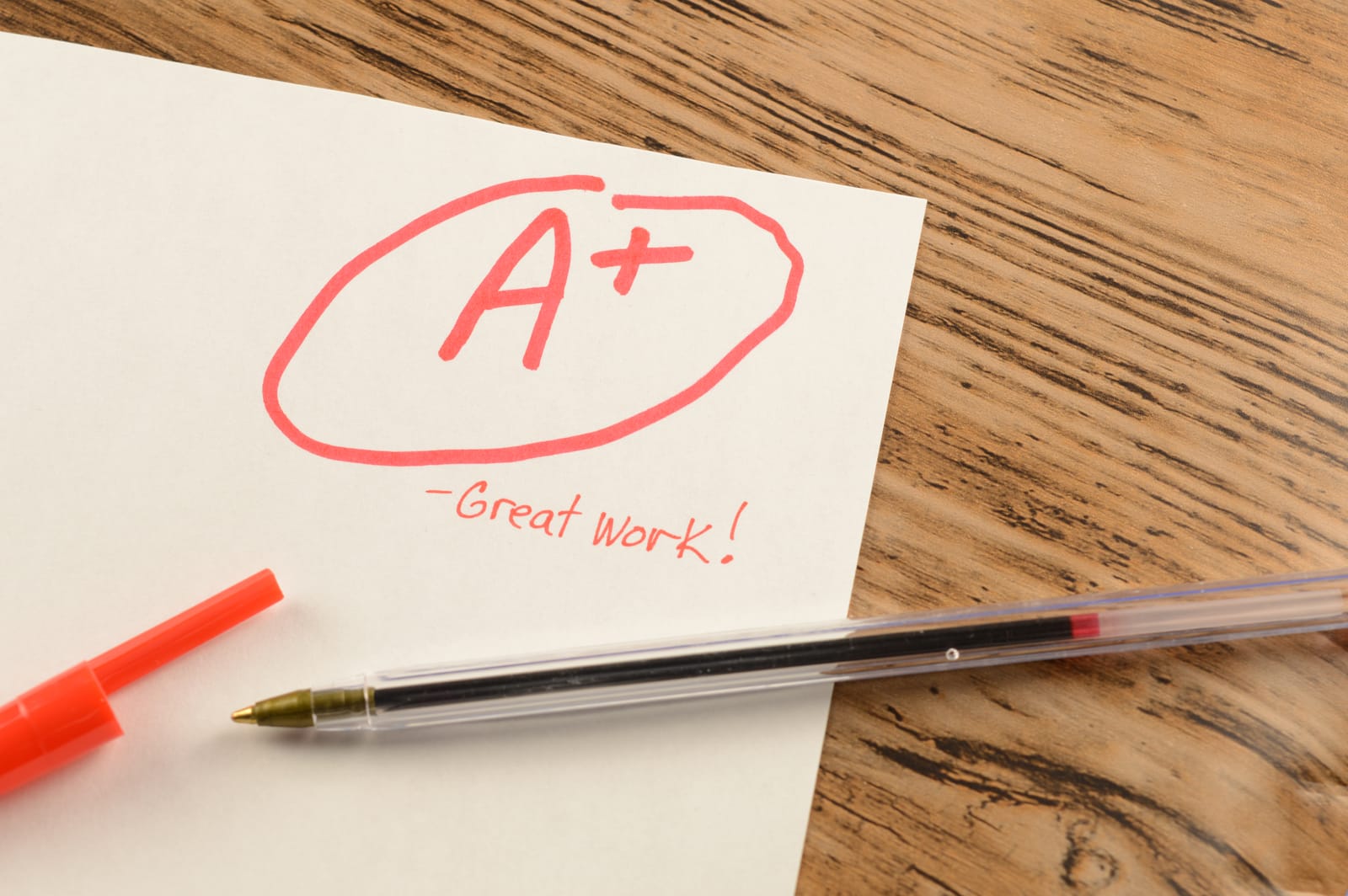 An A+ circled in red marker on a sheet of white paper, with the words Great work! written beneath it and a red ballpoint pen lying across the corner of a wooden desk.