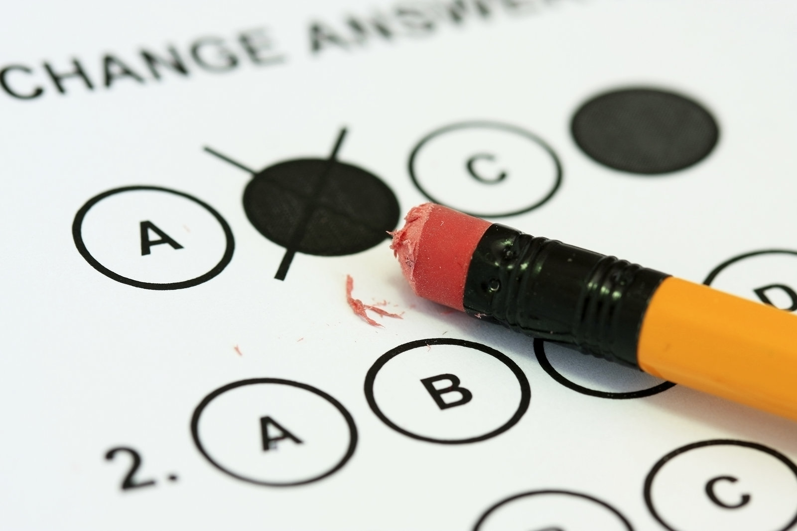 Close-up of a multiple-choice answer sheet headed Change answer, where a pencil eraser is rubbing out a filled-in bubble, leaving eraser crumbs across the row of A, B, C and D options.