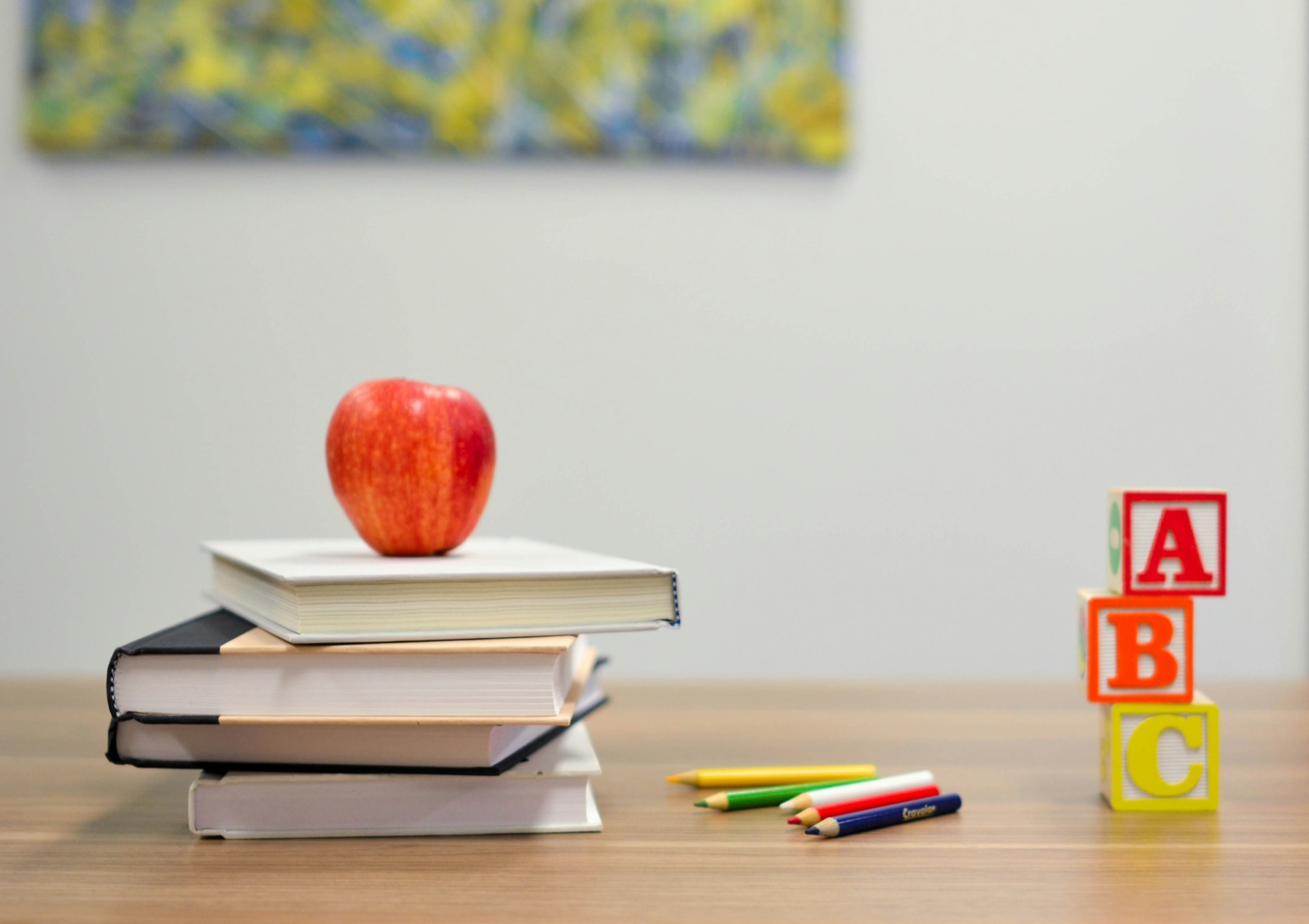 An apple resting on a stack of three hardback books on a wooden desk, coloured pencils beside them and A, B and C alphabet blocks stacked to the right.