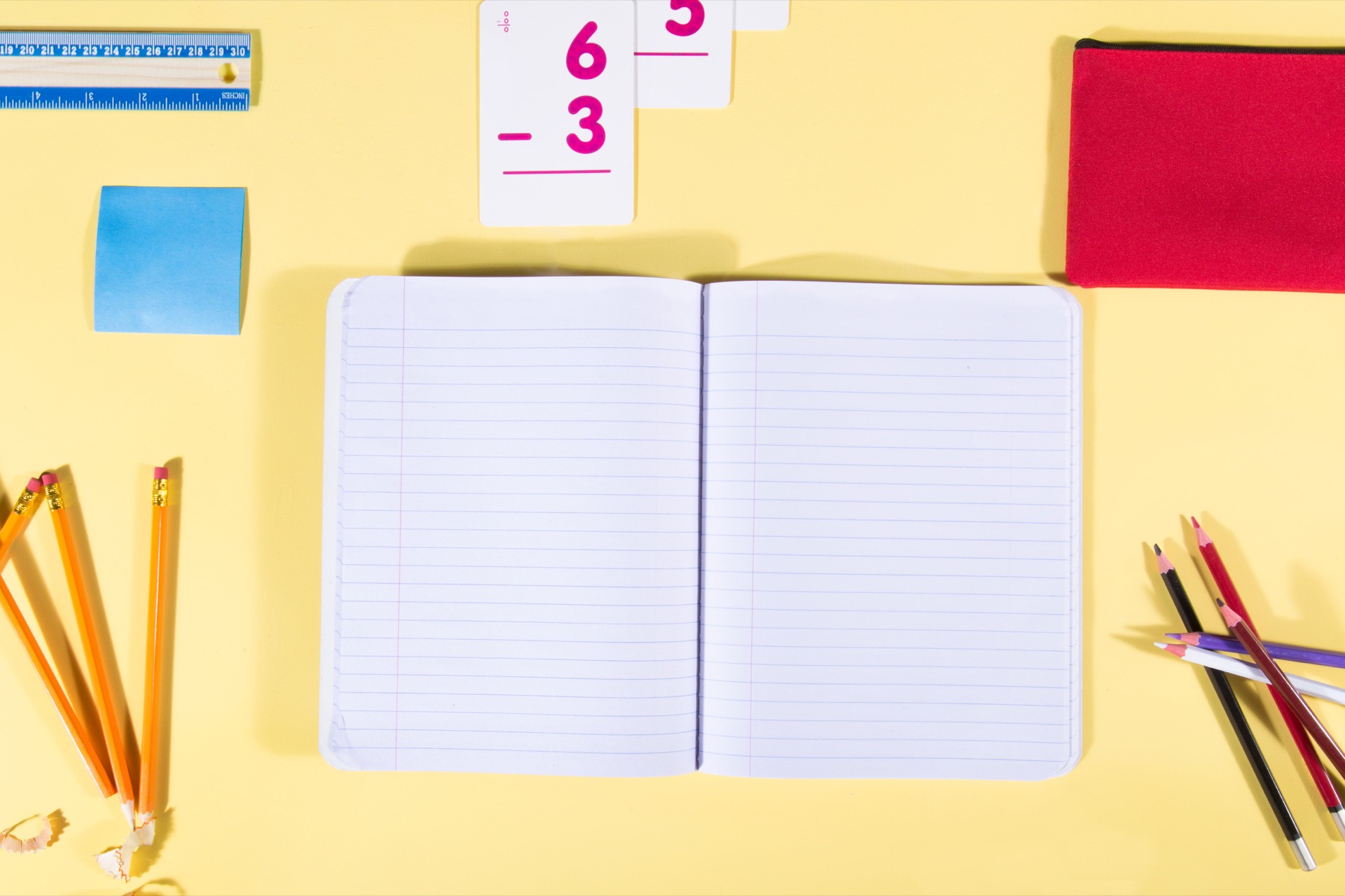 Overhead flat-lay on a yellow background: an open blank exercise book, sharpened pencils with their shavings, coloured pencils, a ruler, a blue sticky note, a red pencil case, and subtraction flashcards reading six minus three.