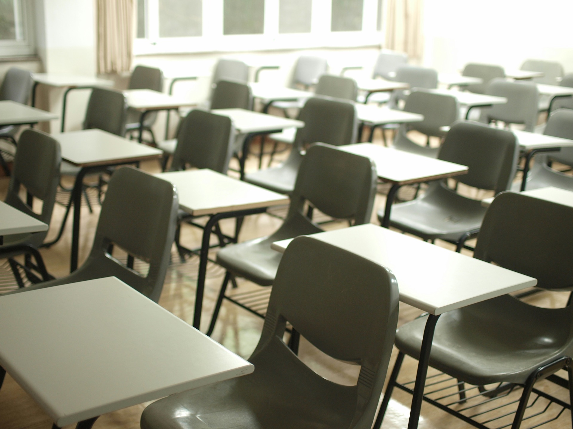 Rows of empty grey chair-desks in an examination room, receding towards a bank of bright windows at the back.